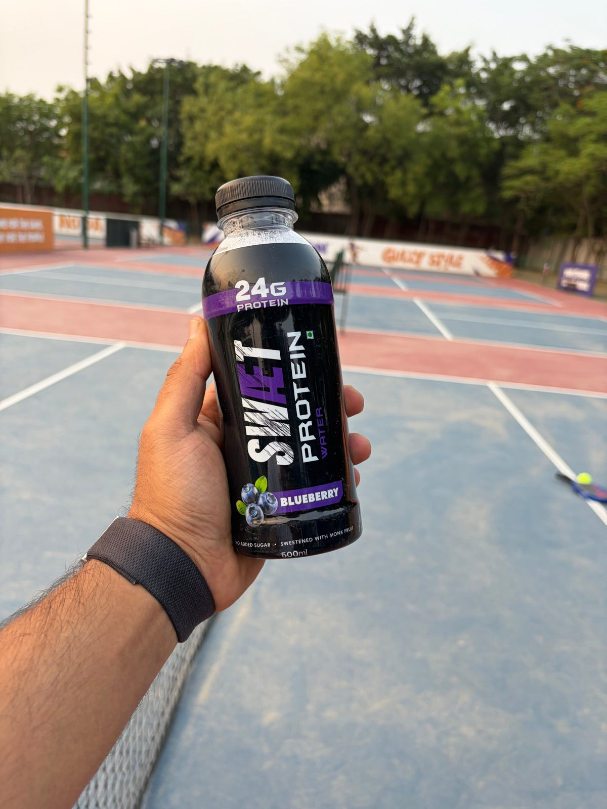 Hand holding SWÆT Blueberry on a tennis court
